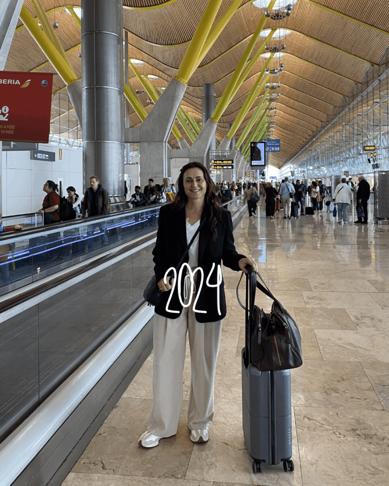 travel_styletips_styleculture_styling_5 airport style photo in Madrid featuring a black blazer, white tee-shirt, cream Camilla and marc pants and sneakers holding a carry-on and wheelie